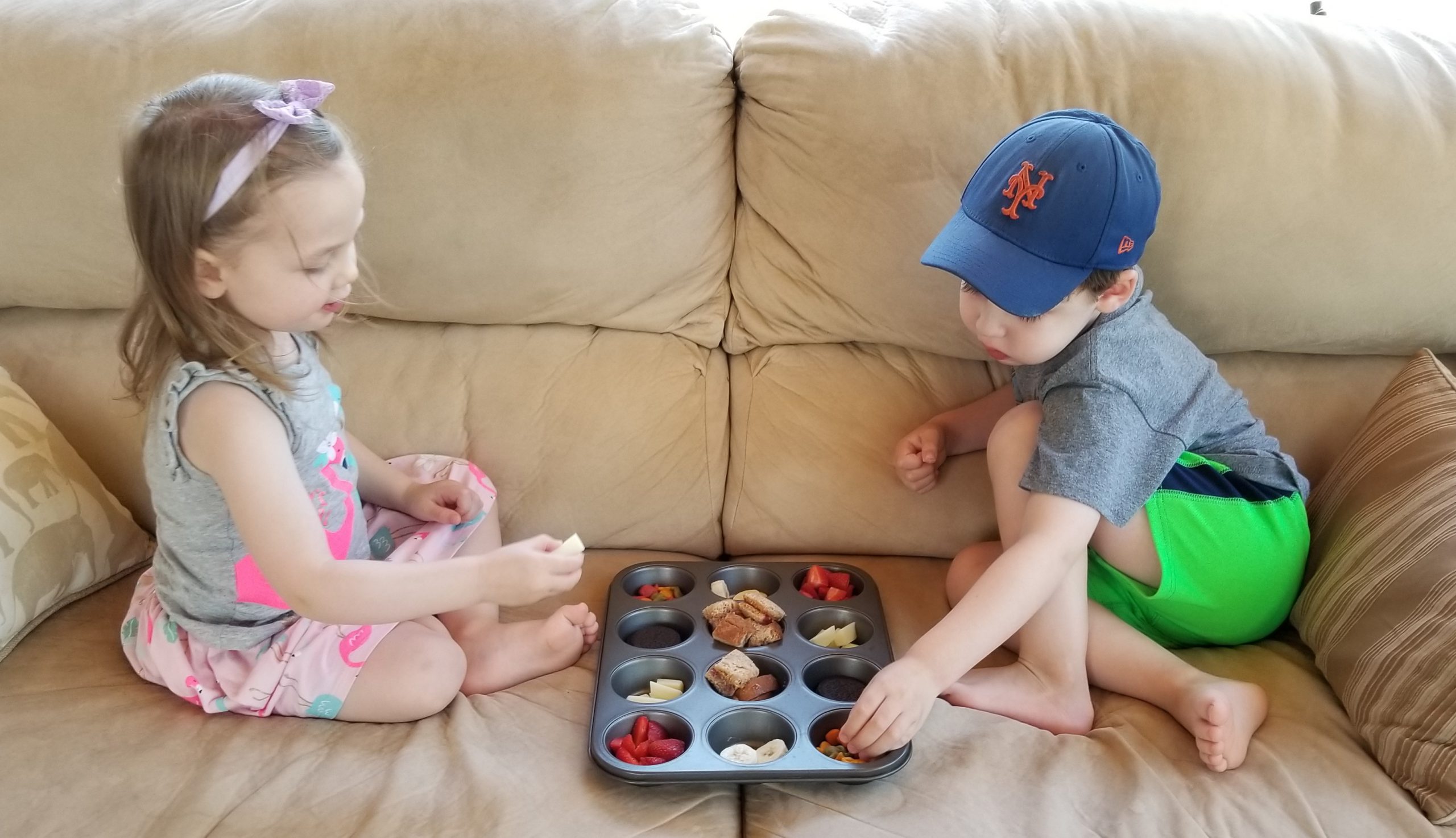 Two young children sit on a couch, a muffin tin filled with snacks between them, and select snacks to taste.