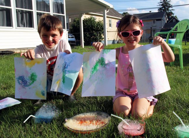 Two children sitting in front of colorful water and holding artwork