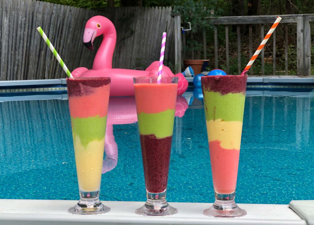 Three multi colored, layered smoothies in pilsner glasses on the side of a pool. A flamingo float floats on the water in the background