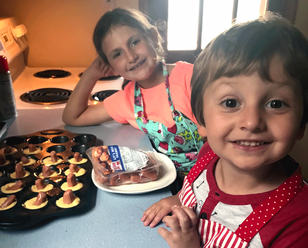 Two children smile at camera with hot dog mini corn muffins next to them on the table