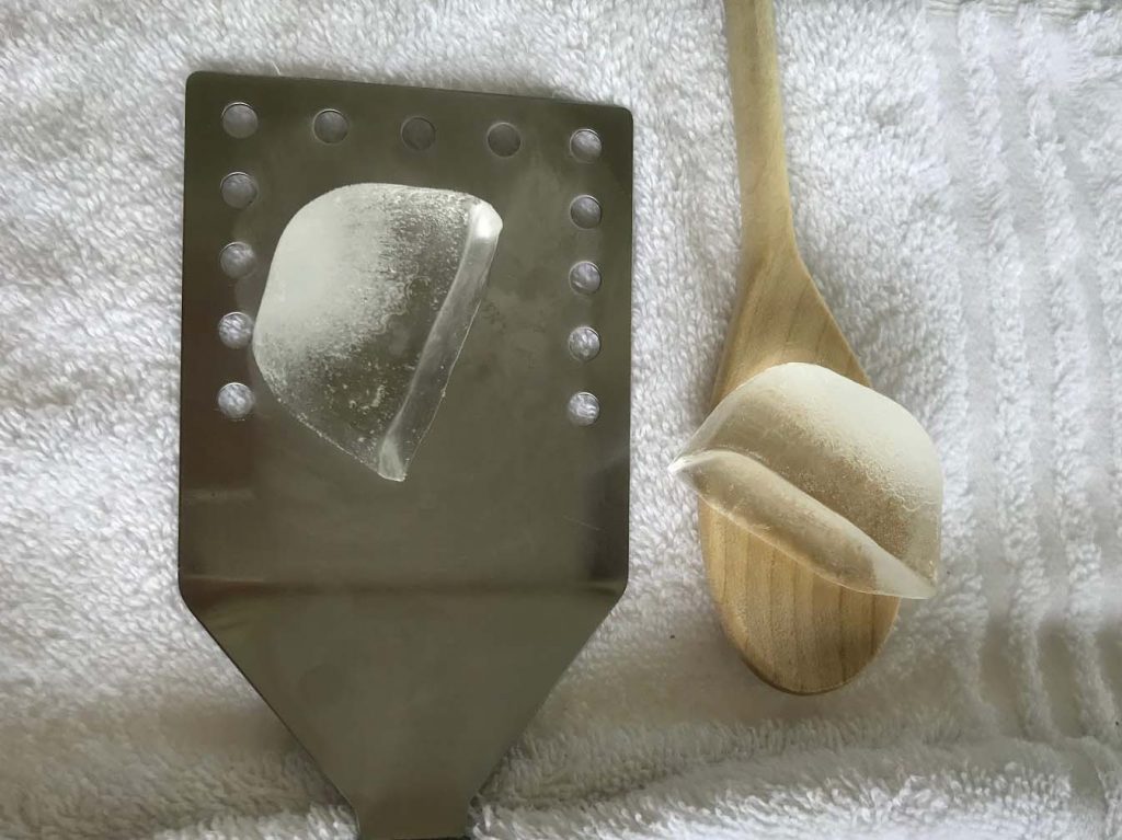 A spatula with an ice cube resting on it next to a wooden spoon, also holding an ice cube