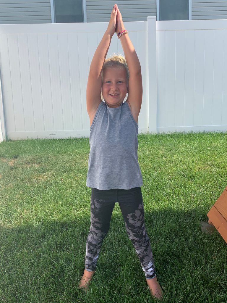 Young girl standing outside in the grass, arms stretched overhead in a yoga pose.