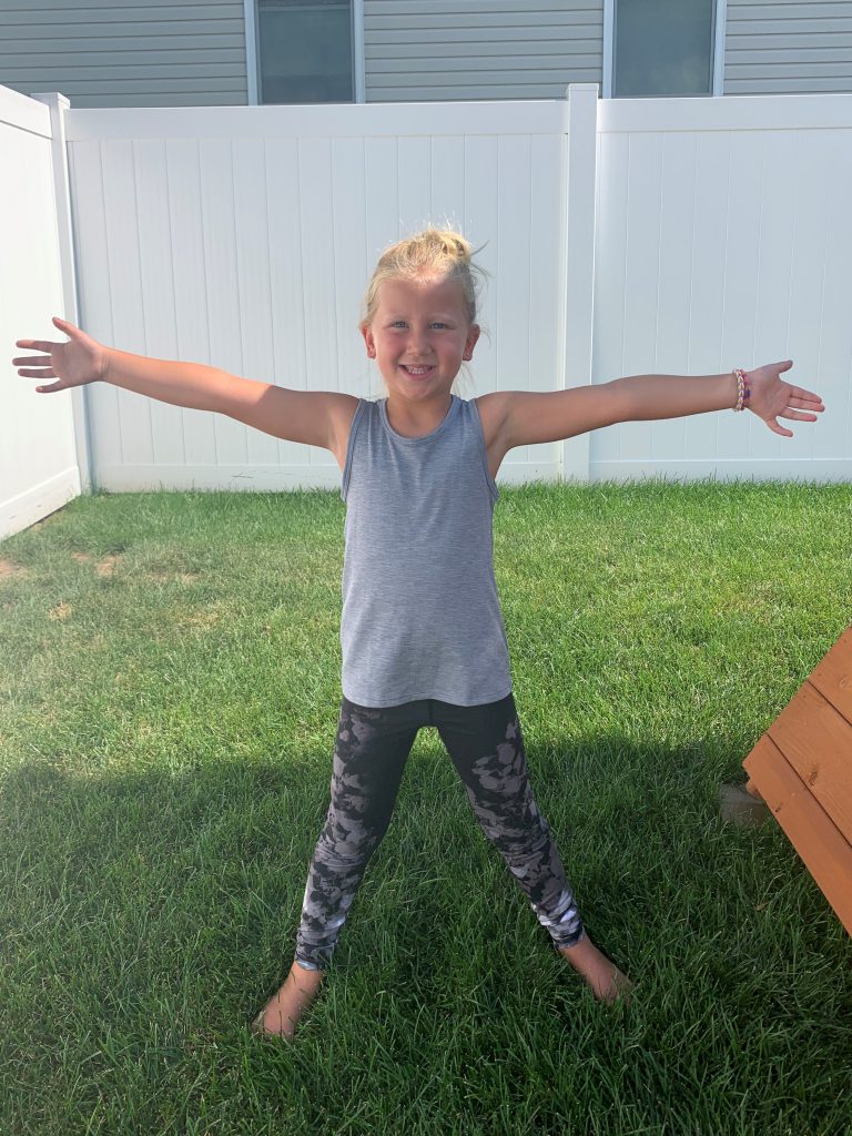 Young girl standing outside in the grass, feet wide and arms outstretched in a yoga pose.