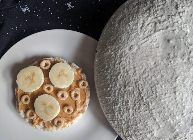 A rice cake covered in peanut butter, cheerios and bananas sits beside a toy moon.