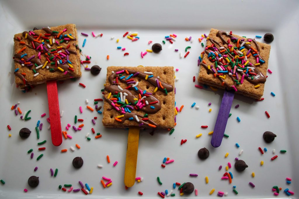 Three S’mores on a Stick rest on a white plate with sprinkles all around them 