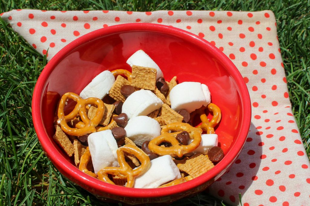 A bowl of S’mores Snack Mix sits on a red and beige polka dot napkin in the grass