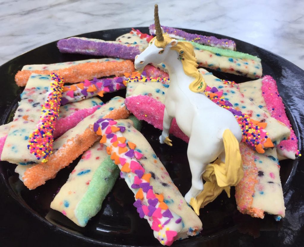A plate of shortbread cookies decorated in bright sprinkles. A figure of a white unicorn is in the center of the plate, surrounded by the cookies.